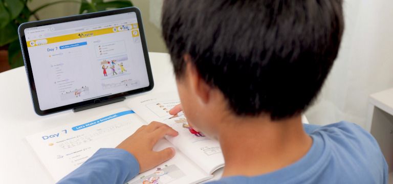 タブレットを見ながらテキストに英語の文字を書こうとしている子供の写真